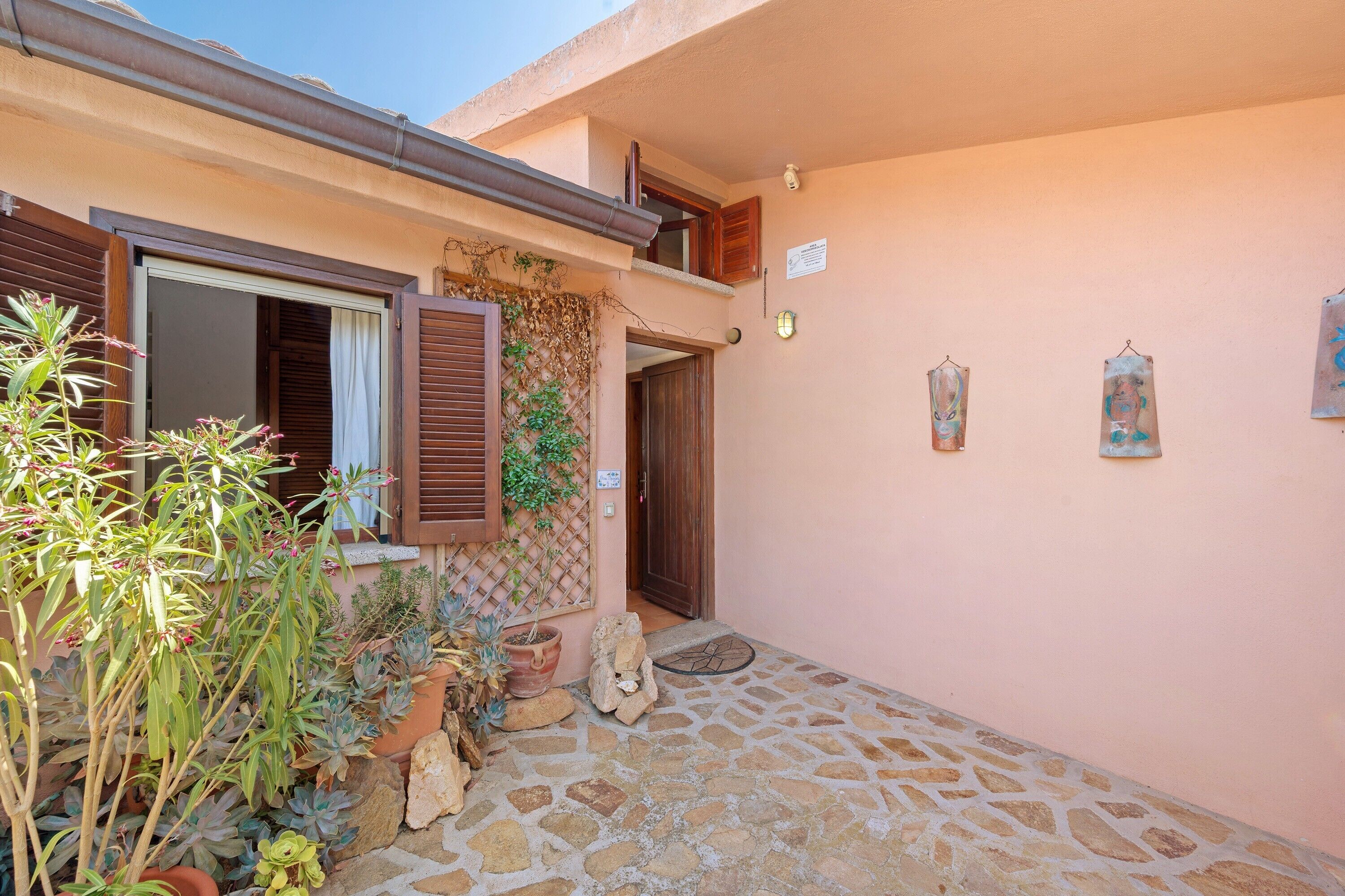 A stone-paved courtyard with a wooden door, a window with shutters, and a wall with a sign and two framed artworks.