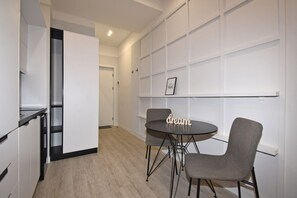 Een moderne keuken met een ronde tafel en twee stoelen, een witte muur met een plank, en een deur naar een andere kamer.