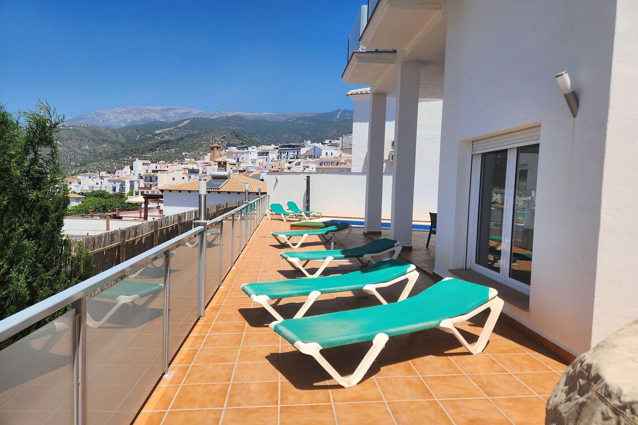 Un balcon avec des fauteuils de jardin verts, une balustrade en verre et une vue sur une ville et des montagnes.