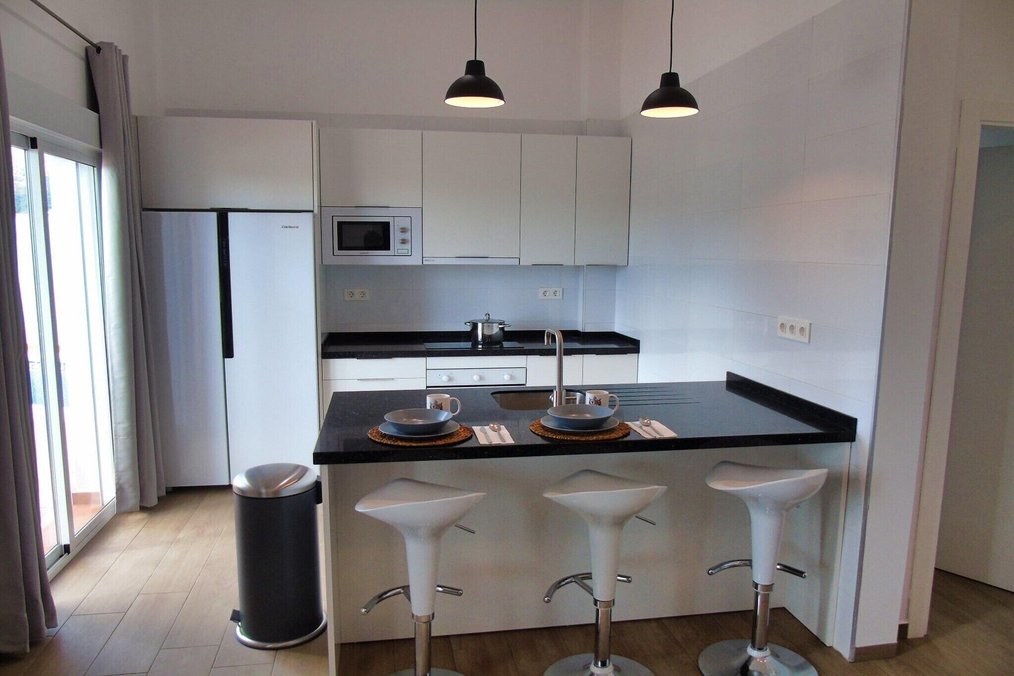 Una cocina moderna con armarios blancos, encimera negra, microondas y lavamanos. Hay tres taburetes blancos de bar y dos lámparas colgantes.