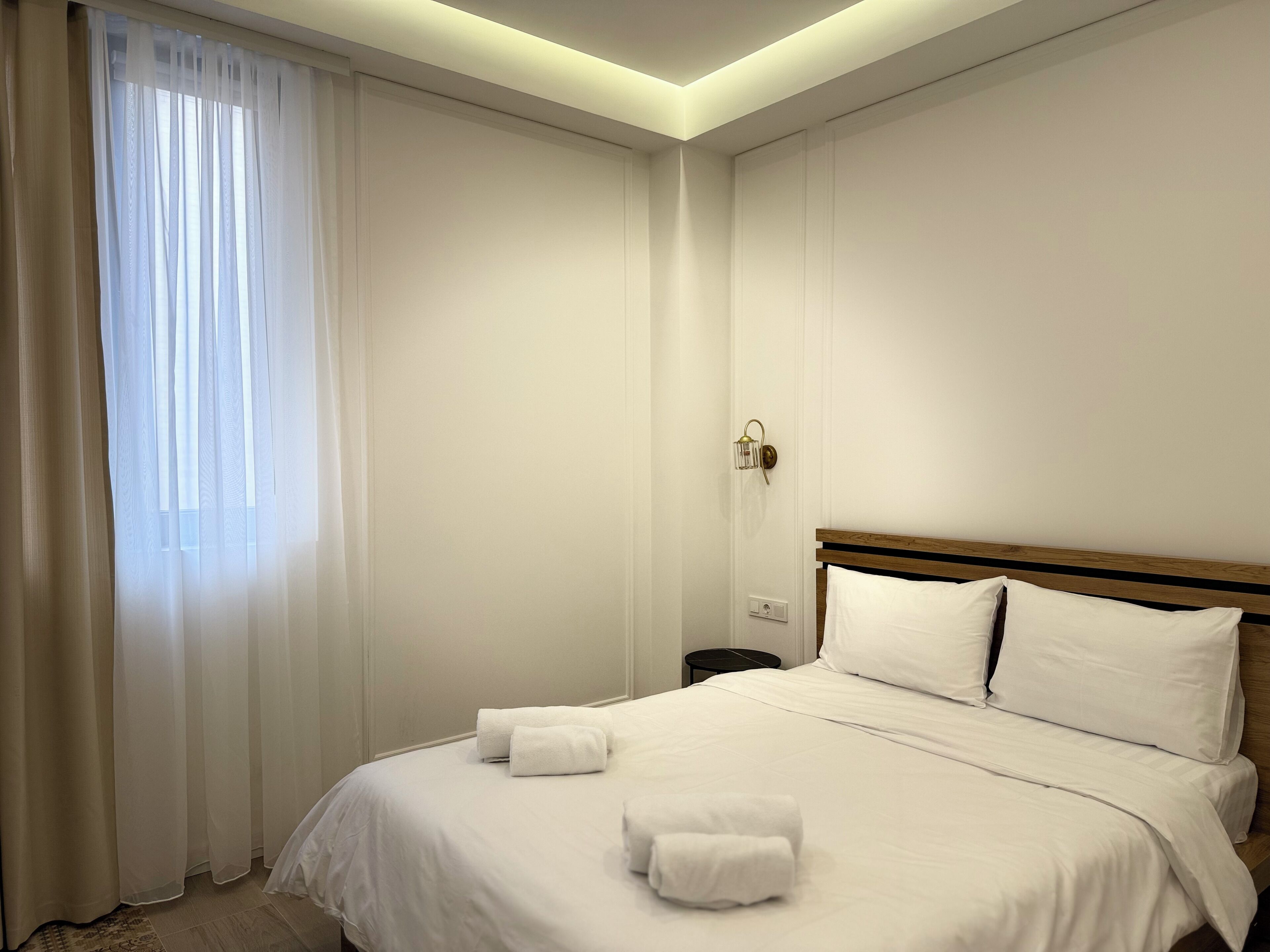 Une chambre d’hôtel avec un lit bien fait, une table de chevet, une lampe et une fenêtre ornée de rideaux légers.