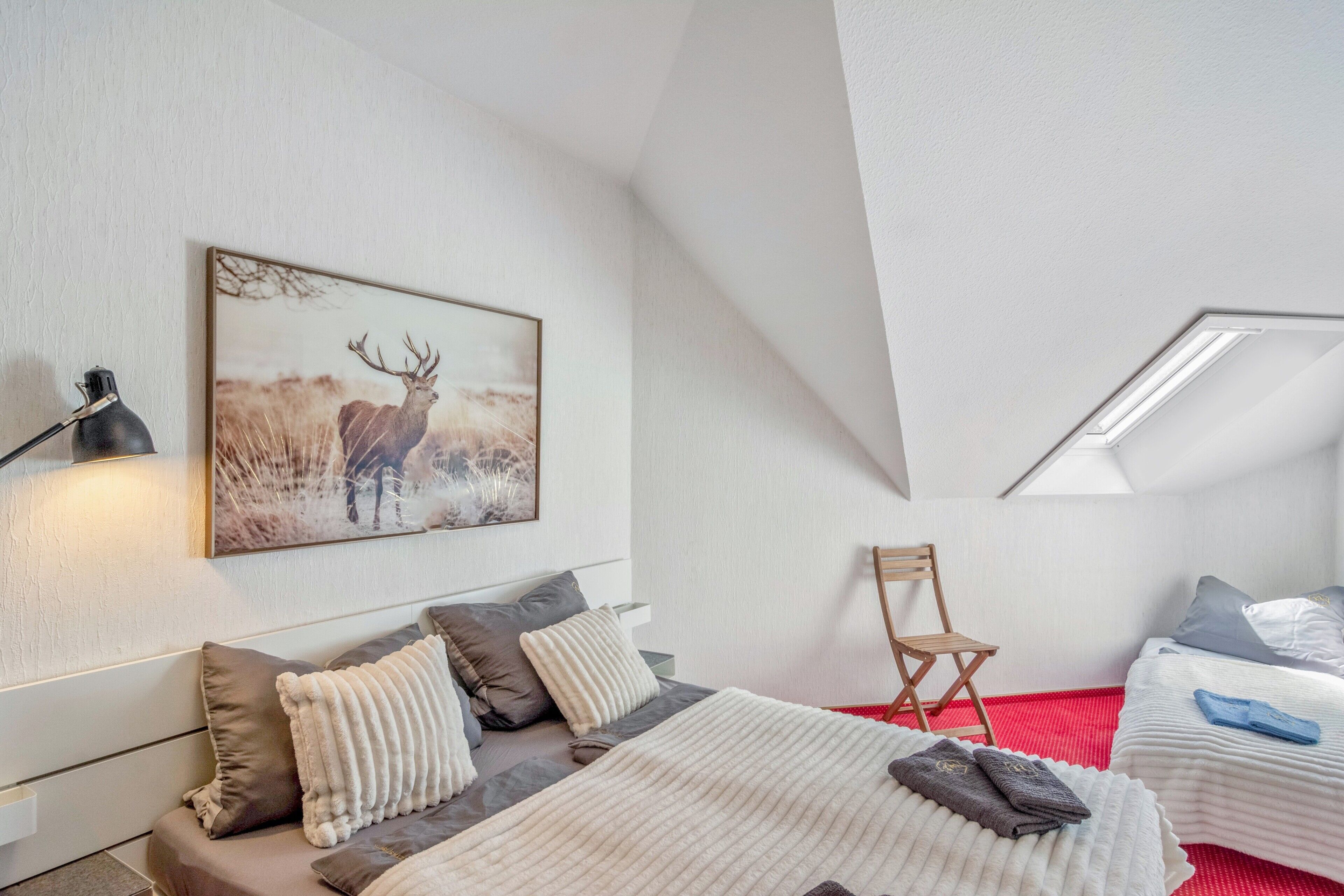 Ein Schlafzimmer mit einem Bett, Kissen, einem Sessel, einem Schreibtisch, einer Lampe und einem Bild von einem Reh.