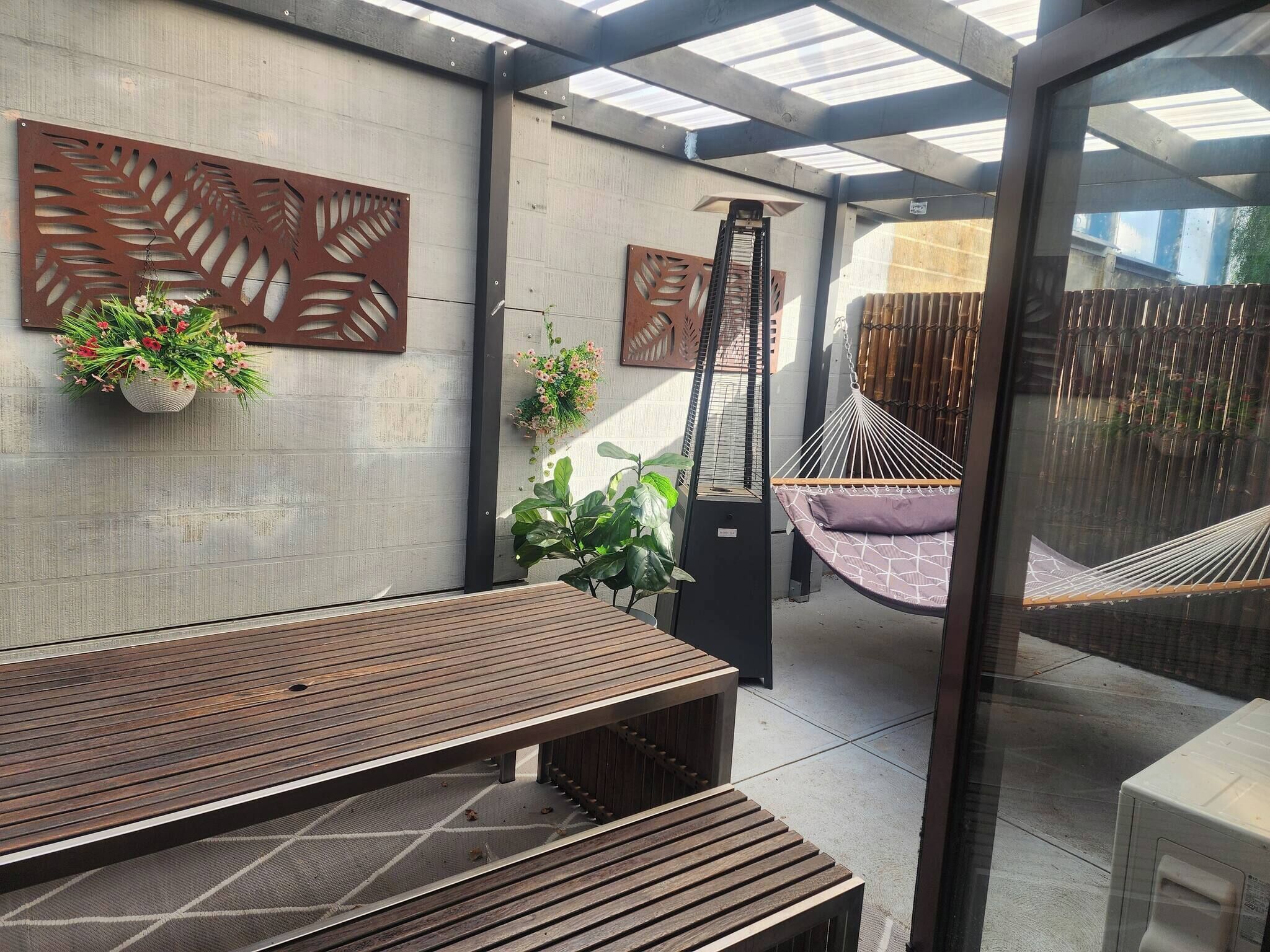 A covered patio with a wooden bench, decorative wall panels, hanging plants, and a hammock.