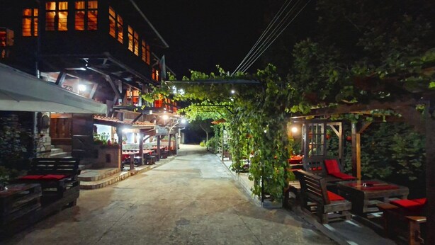 Calle iluminada de noche con edificios, una marquesina y mesas al aire libre.