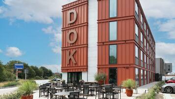 A multi-story building with a prominent 'DOK' sign on the facade, outdoor seating area with tables and chairs, and a clear sky in the background.