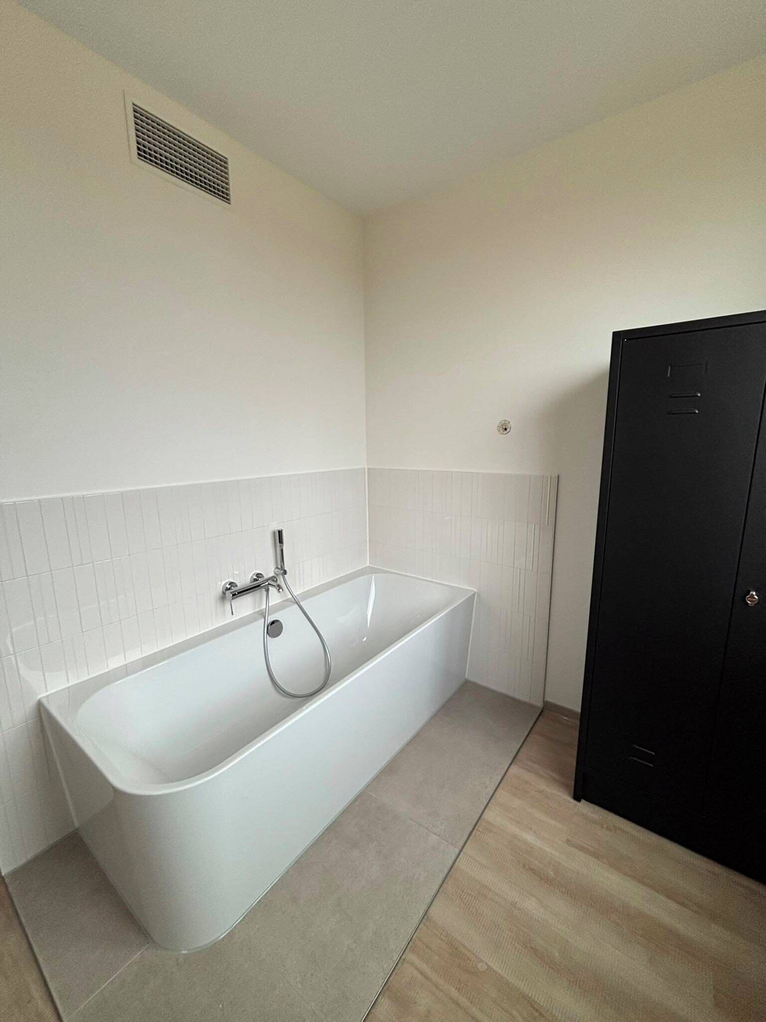 A modern bathroom with a white bathtub, a black wardrobe, and a vent on the wall.