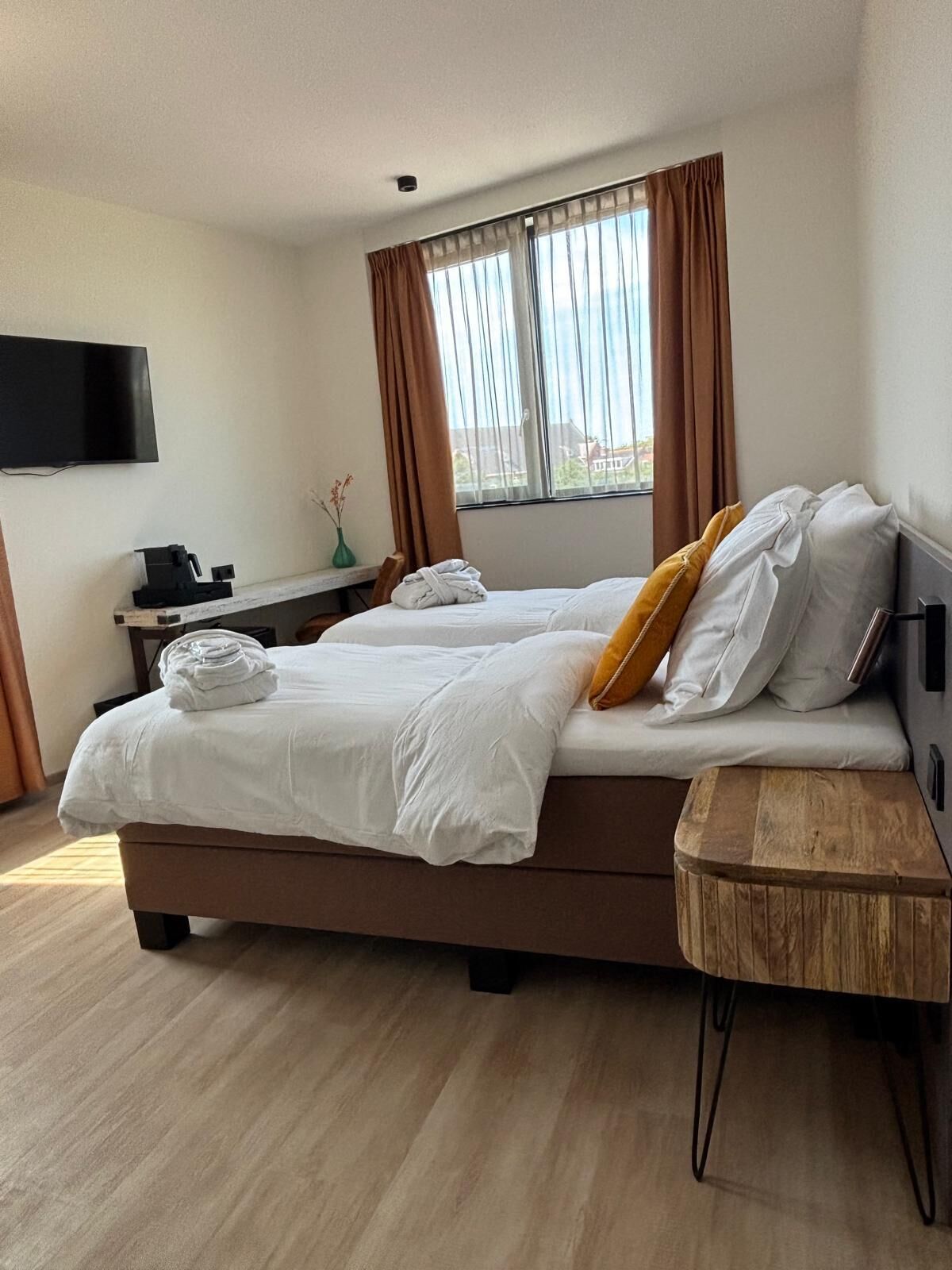 A hotel room with a bed, a small wooden side table, a desk with a chair, a television mounted on the wall, and a window with curtains.
