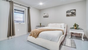 Un dormitorio con cama, ventana con cortinas, mesita de noche con una planta y dos cuadros en la pared.
