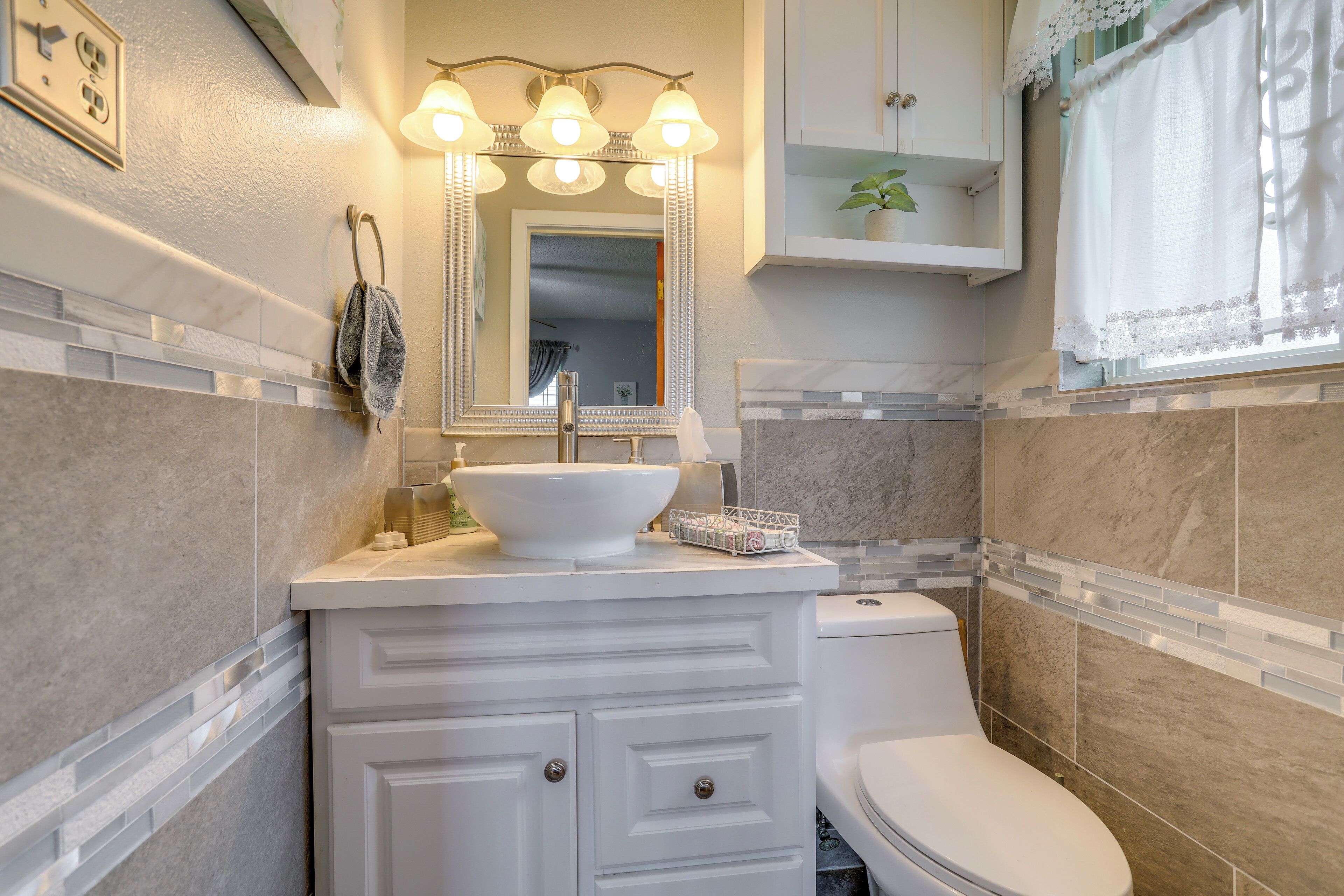 A bathroom with a white sink, vanity, and toilet. There is a mirror with lights above it, and a towel hanging on the wall.
