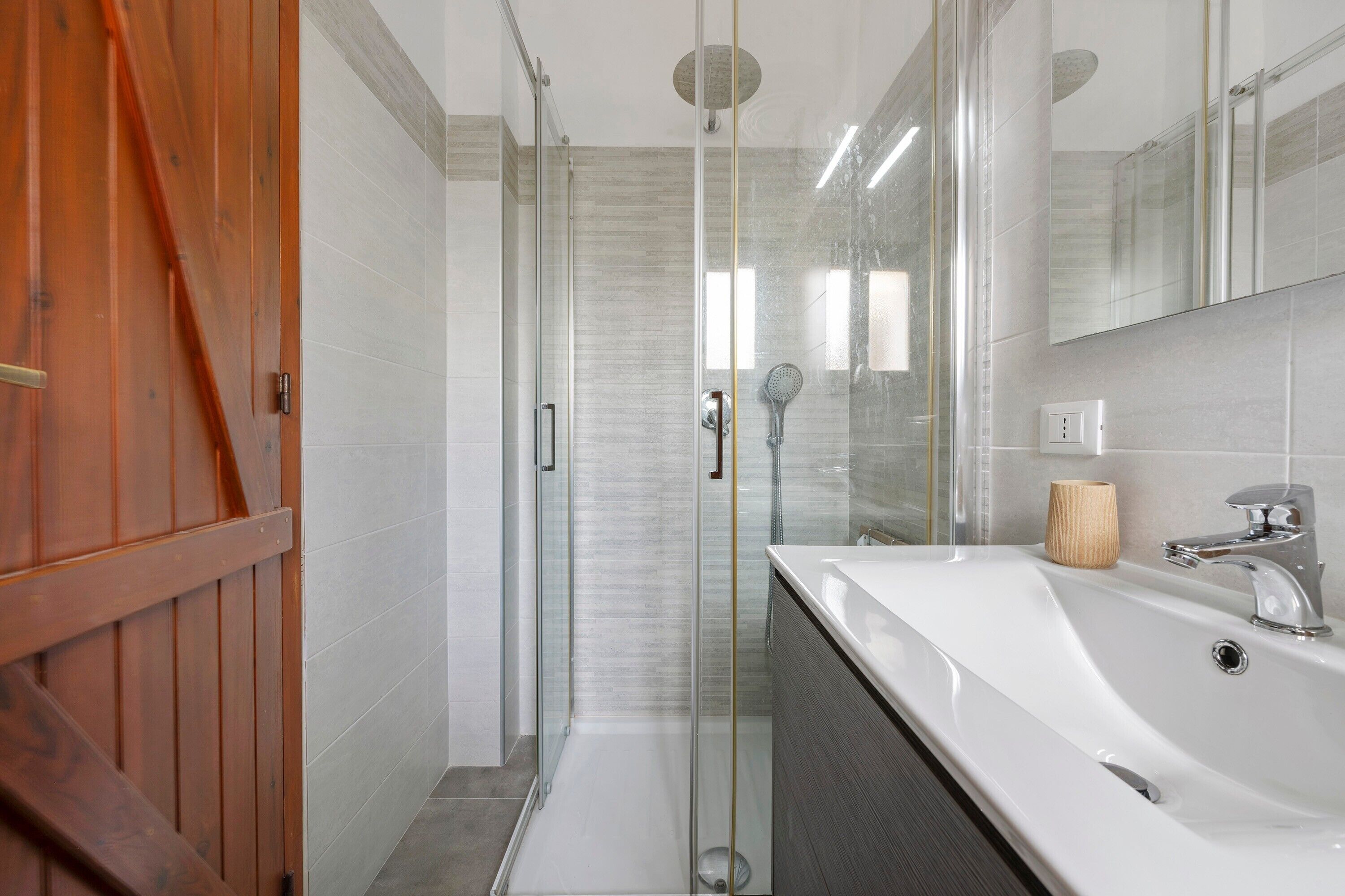 A modern bathroom with a glass shower enclosure, a wooden door, and a white sink with a wooden bowl.