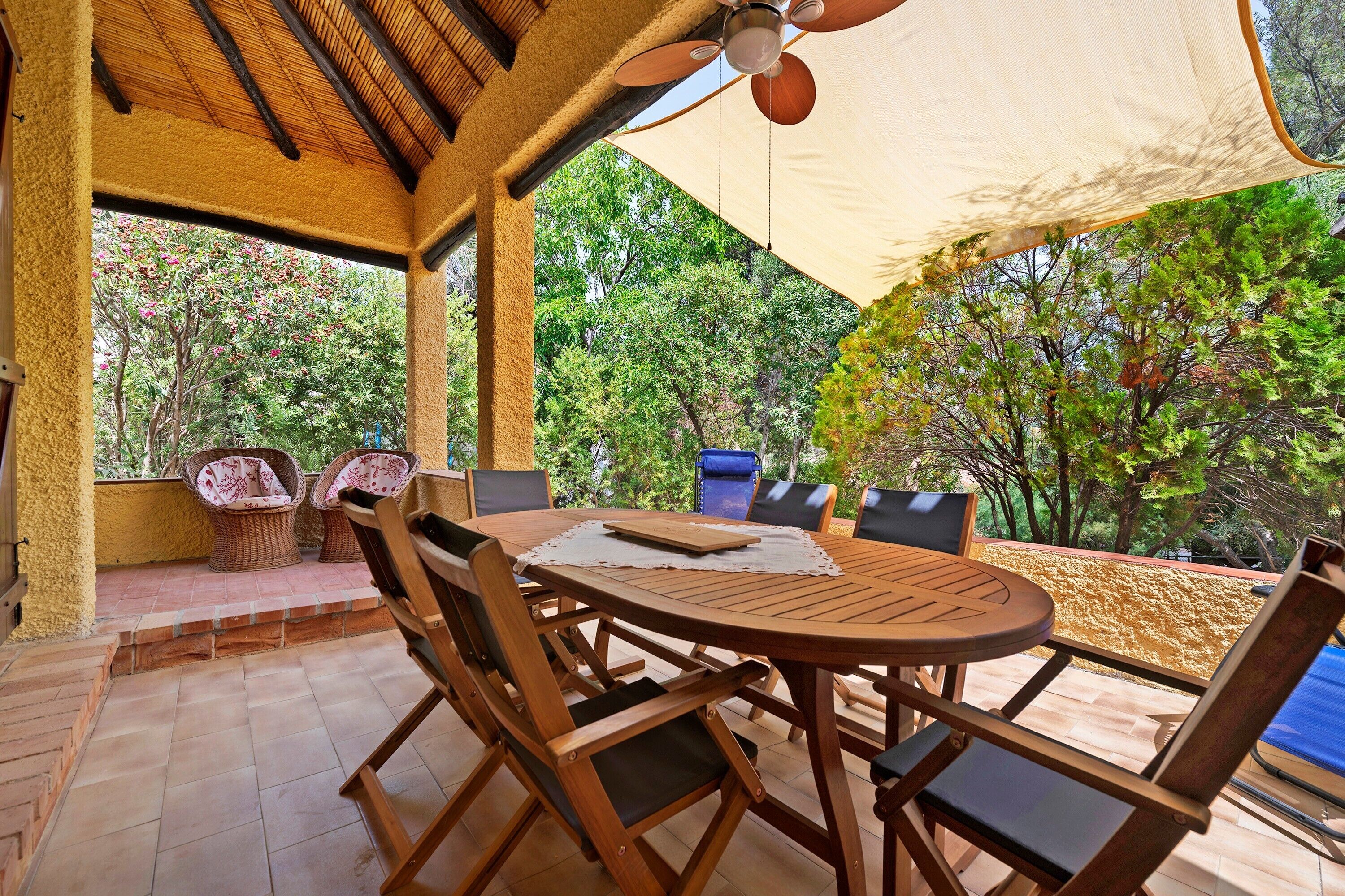 Eine Terrasse im Freien mit einem runden Holztisch und Stühlen, einem Deckenventilator und einem großen Sonnensegel, das Schatten spendet.