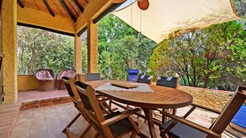Eine Terrasse im Freien mit einem runden Holztisch und Stühlen, einem Deckenventilator und einem großen Sonnensegel, das Schatten spendet.