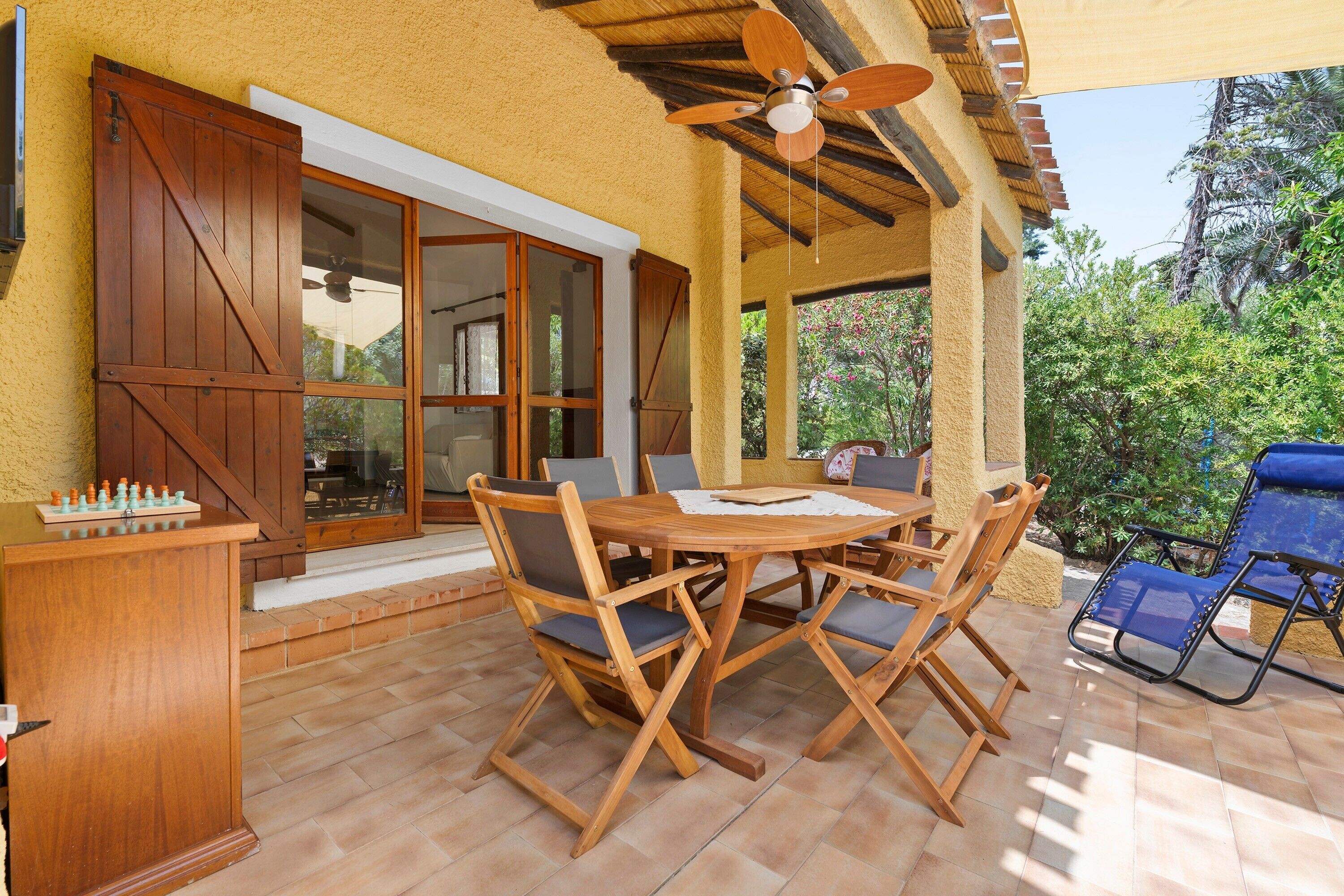 An outdoor patio with a wooden table and chairs, a ceiling fan, and a view of greenery.