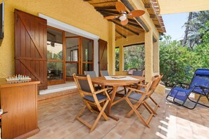 Um pátio externo com mesa e cadeiras de madeira, um ventilador de teto e vista para o verde.