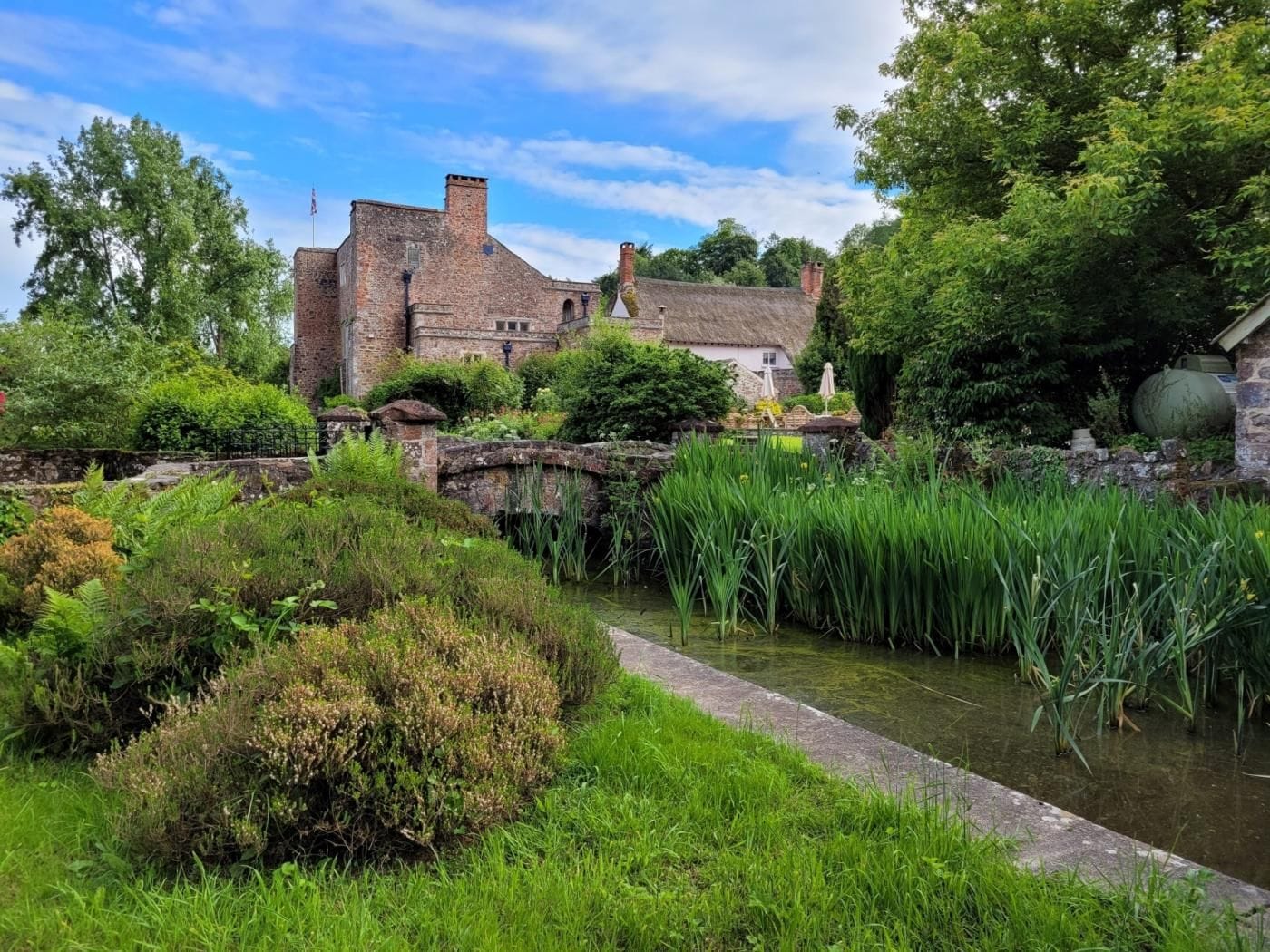 Een historisch gebouw met rieten dak, omgeven door weelderige begroeiing en een klein waterkanaal.