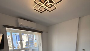Une chambre à coucher comprenant un lit, une commode en bois, un bureau et une fenêtre donnant sur des immeubles.