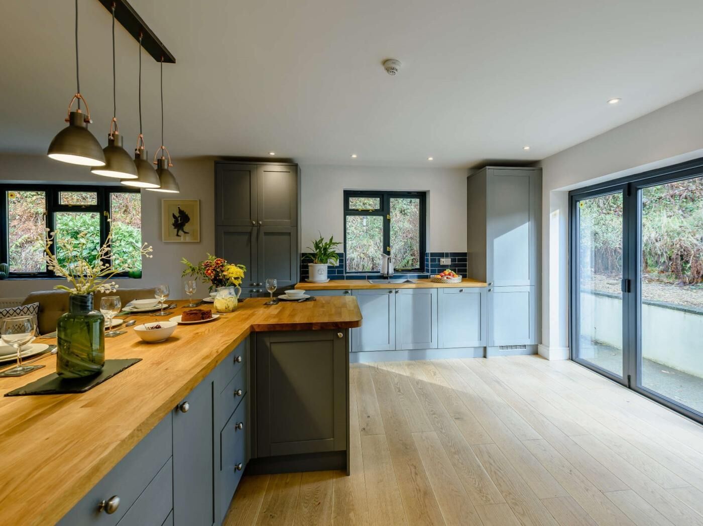 A modern kitchen with a wooden countertop, grey cabinets, pendant lights, and large windows.