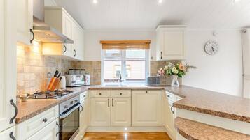 Une cuisine avec des meubles blancs, un plan de travail en bois, une cuisinière, un évier et une fenêtre avec des stores.
