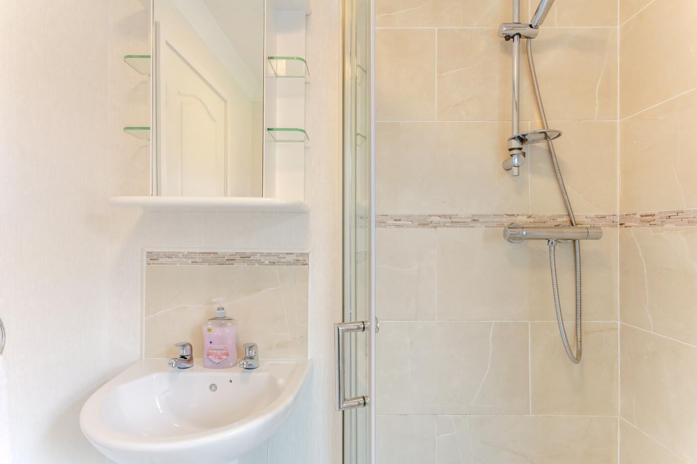 Un bany amb dutxa de vidre, un lavabo blanc i un mirall amb una prestatgeria a sobre.