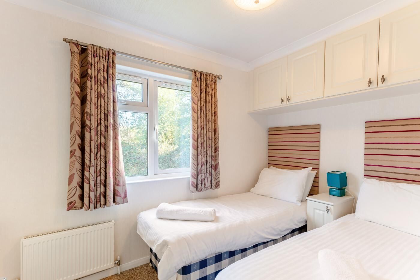 A bedroom with two beds, a window with curtains, a radiator, and a nightstand with a lamp.