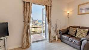 Un salon avec un canapé en cuir marron, un téléviseur, un lustre et une vue sur la mer, visible à travers une grande porte vitrée.
