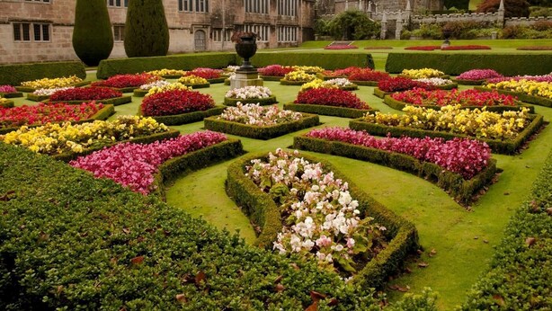 Ein formeller Garten mit geometrisch angelegten Blumenbeeten, einer zentralen Fontäne und einem historischen Gebäude im Hintergrund.