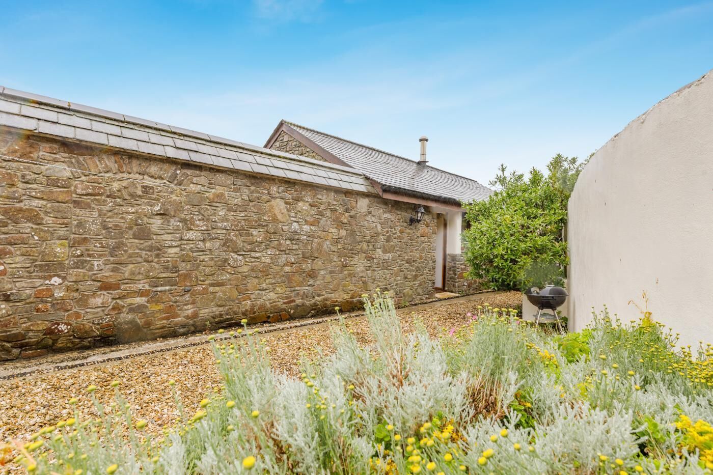 Un bâtiment en pierre avec un toit de chaume, un chemin de gravier et un barbecue en premier plan.