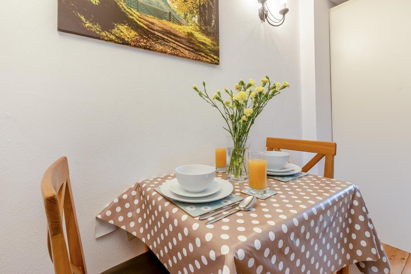 A dining area with a table set for two, featuring a polka dot tablecloth, white plates, a vase with flowers, and glasses of orange juice.
