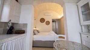 A room with a bed, a small table, a chair, a cabinet, and a kitchen area with a coffee machine. - Trullo NiDo – Entire Property for Exclusive Use (Latiano)