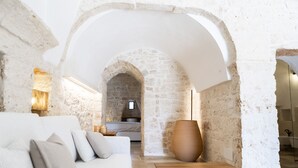A room with a white sofa, a wooden table, a large vase, and a stone wall with an arched doorway leading to a kitchen area. - Trullo NiDo – Entire Property for Exclusive Use (Latiano)