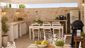 An outdoor kitchen with a stone wall, white furniture, and a grill. - Trullo NiDo – Entire Property for Exclusive Use (Latiano)