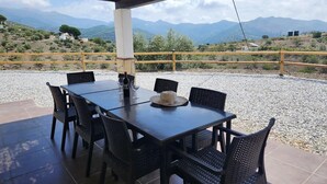 Uma área de refeições ao ar livre coberta, com uma mesa e cadeiras, um chapéu sobre a mesa e vista para montanhas ao fundo.