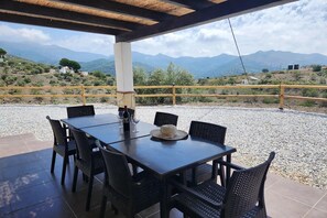Ein überdachter Essbereich im Freien mit Tisch und Stühlen, ein Hut auf dem Tisch und Blick auf Berge im Hintergrund.