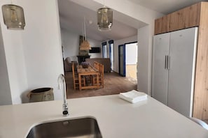 Une cuisine moderne avec un plan de travail blanc, un évier en acier inoxydable et des armoires en bois. On y trouve un coin repas avec une table et des chaises en bois, et une vue sur l’extérieur grâce à une porte vitrée.