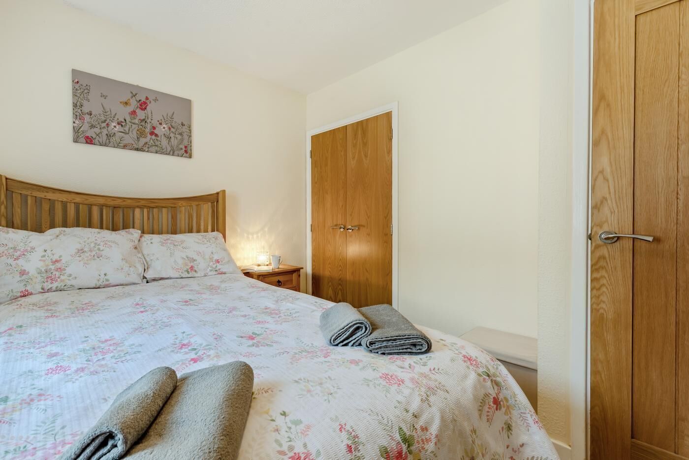 A bedroom with a wooden headboard, a bed with floral bedding, a nightstand with a lamp, and a wooden wardrobe.