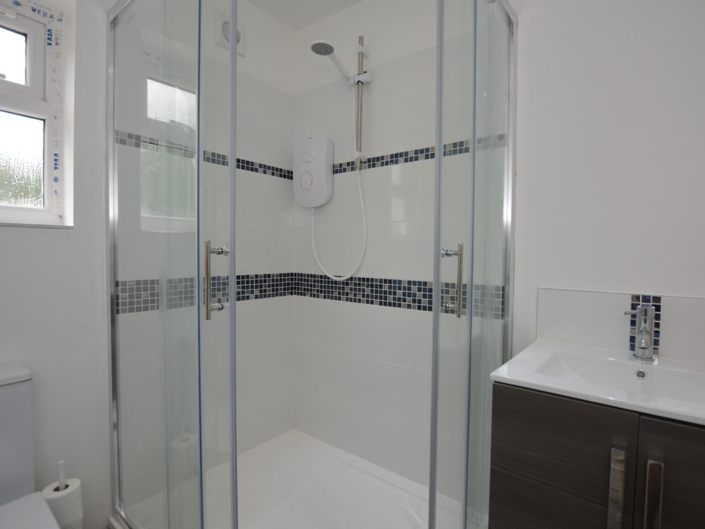 A glass-enclosed shower with a handheld showerhead, a white sink, and a tiled shower wall.