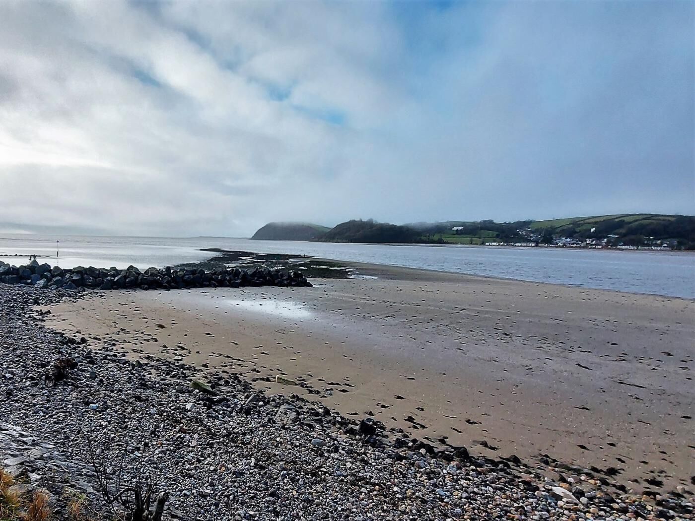 A pebbly beach with a rocky breakwater, calm sea, and distant hills.