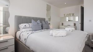 Ein ordentlich bezogenes Bett mit Kopfteil, ein Nachttisch, ein Spiegel und eine Wand mit der Aufschrift 'LOVE'.