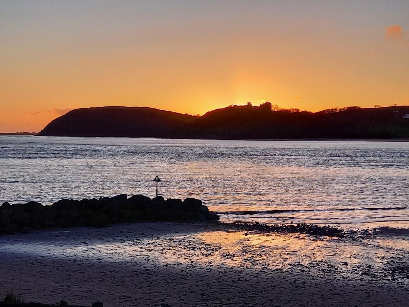Een zonsondergang boven het water met een rotsachtige kustlijn en een heuvel in de verte.