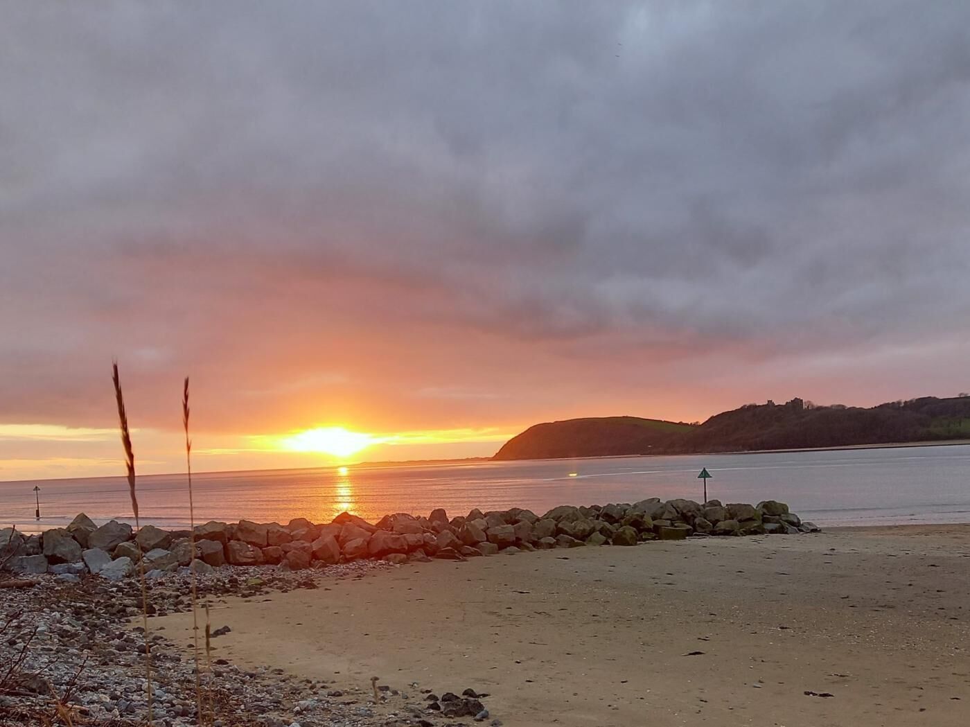 A beach with a sunset, rocky shoreline, and a distant hill.