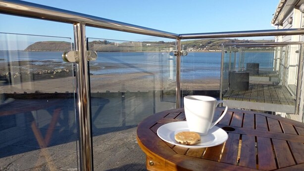 Eine Terrasse mit Glasgeländer, einem Holztisch, einer weißen Tasse und einem Keks.