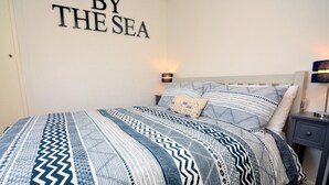 A bedroom with a bed, a bedside table, a nightstand, and a wall sign that reads 'BY THE SEA'. - 3 Bed in Bideford (oc-55363) (Appledore)