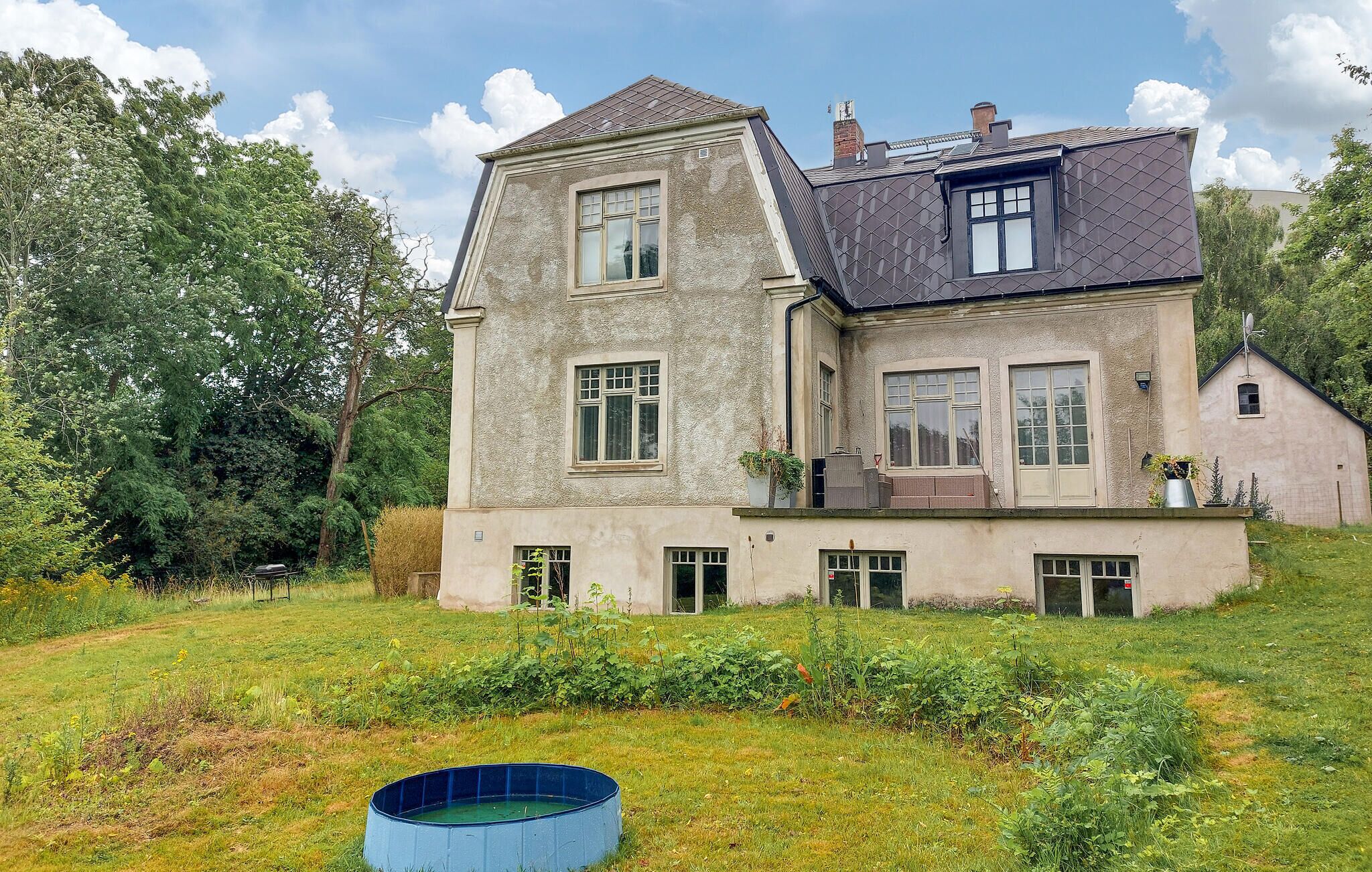 Une maison à deux étages, dotée d’un toit en pente, de nombreuses fenêtres et d’un petit jardin avec une piscine bleue.