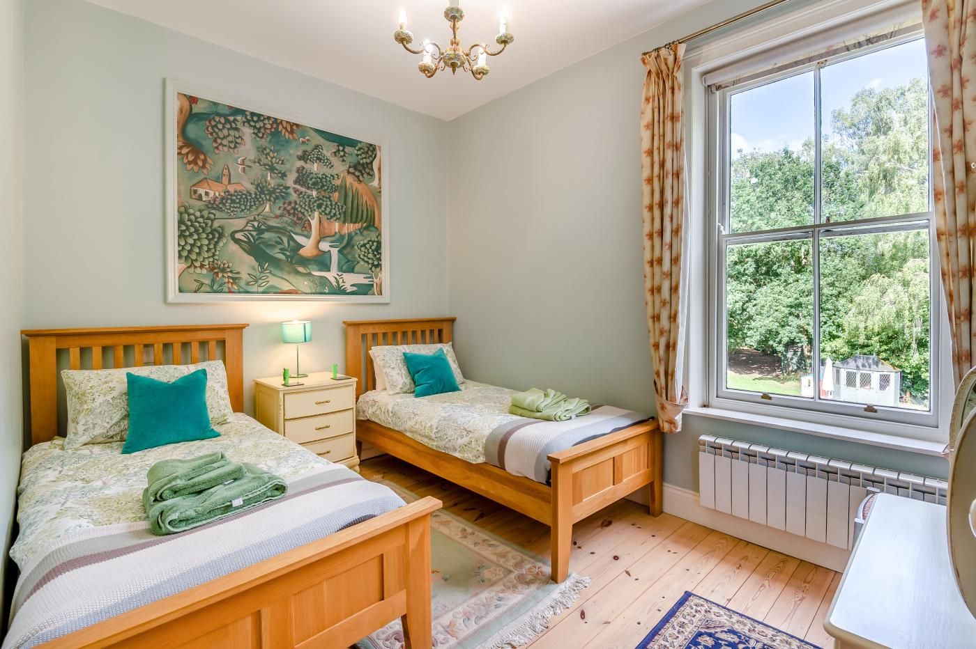 A bedroom with two wooden beds, a nightstand, a painting, and a window with a view of greenery.