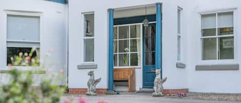 Un bâtiment blanc avec une porte et des fenêtres bleues, un banc en pierre et deux sculptures d’animaux en pierre.