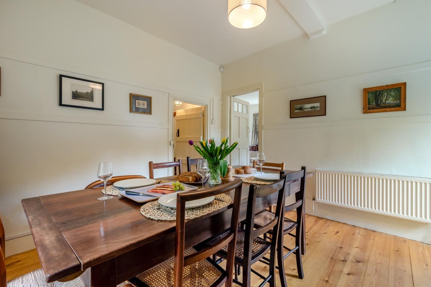A dining area with a wooden table, chairs, and a vase of flowers. There are framed pictures on the walls and a radiator on the floor.