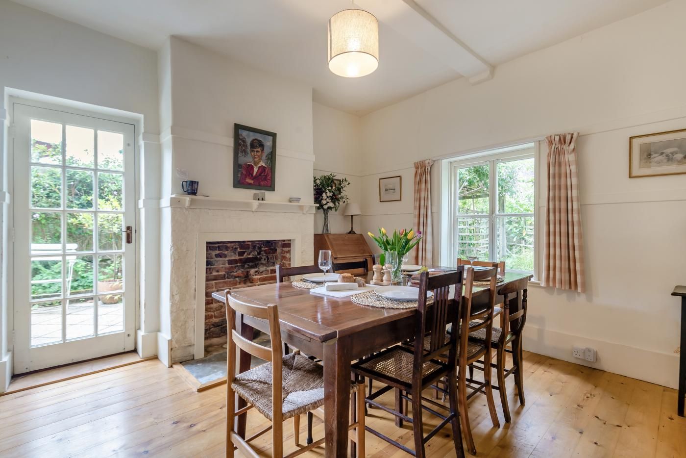 A dining room with a brick fireplace, wooden dining table, and chairs. There is a window with curtains and a painting on the wall.