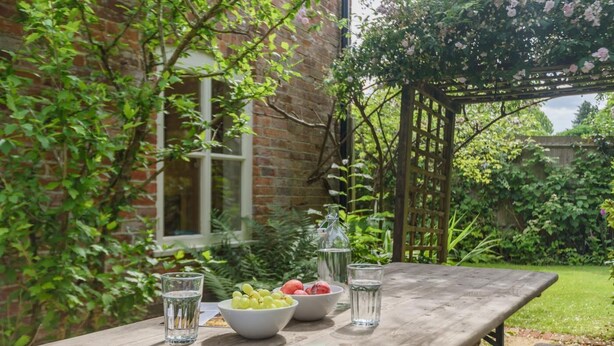 A wooden table with a bowl of fruit, a bottle, and glasses, set against a brick wall and greenery. - 4 Bed in Lyndhurst (oc-57407) (Lyndhurst)