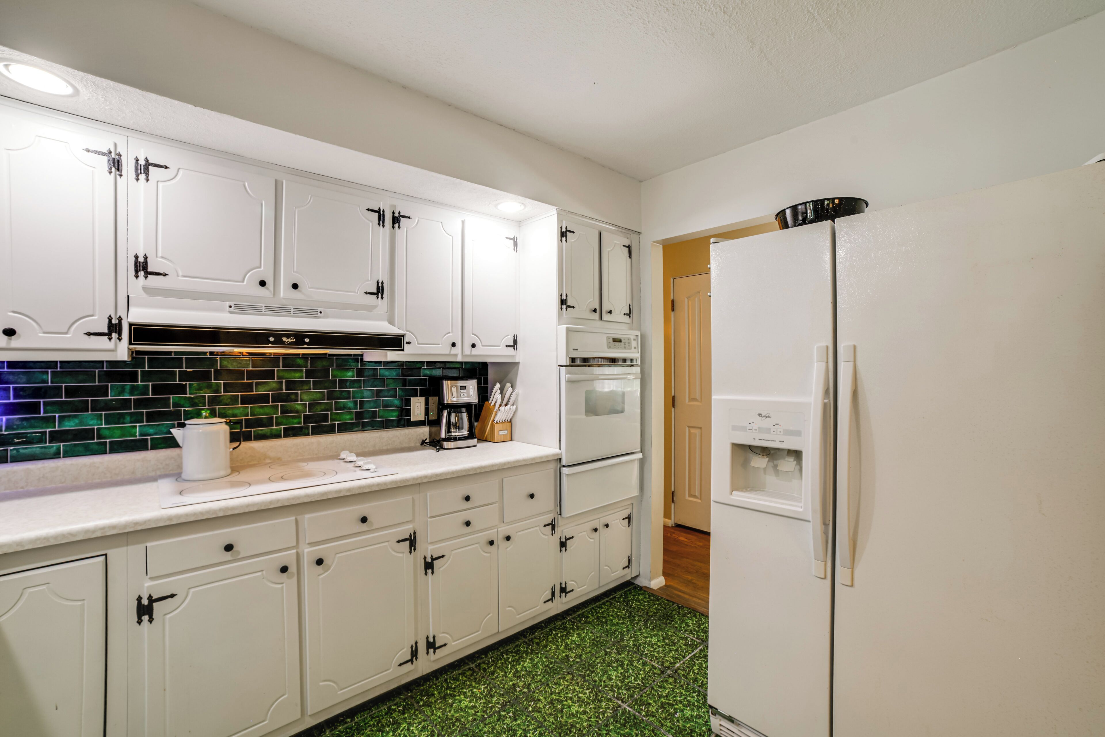 Una cocina con armarios blancos, salpicadero de azulejos verdes, cafetera y refrigerador.
