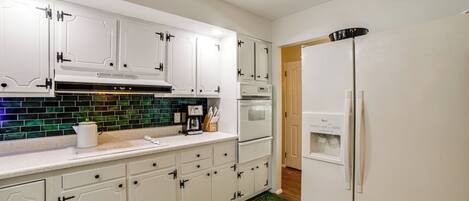 Una cocina con armarios blancos, salpicadero de azulejos verdes, cafetera y refrigerador.